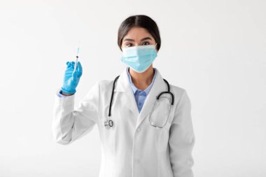 Eldiven ve koruyucu maske takan, tıp üniformalı genç Hintli kadın aşılı bir şırınga sergiliyor. Beyaz bir geçmişe karşı duruyor, toplumundaki sağlık girişimlerine hazır..