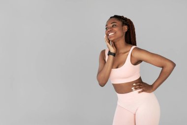 Spor kıyafetli genç siyahi kadın spor takip cihazı takarken gülümseyerek yana bakıyor. Gri arka plan aktif yaşam tarzını ve kendine güvenen duruşunu vurguluyor..
