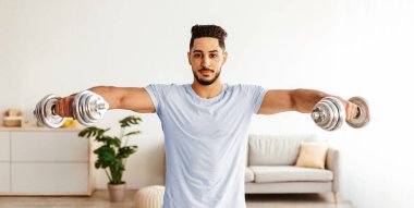 Bir adam iyi aydınlatılmış oturma odasında dambıllarla omuz kaldırır. Odada rahat bir kanepe ve bitki var. Egzersiz için rahat bir atmosfer yaratıyor..