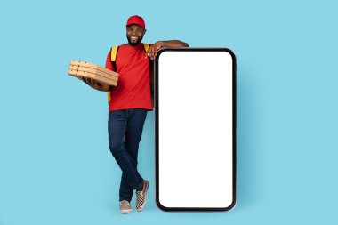 Delivery Service App. Black Courier Man With Pizza Boxes In Hand Standing Near Big Blank Smartphone With White Screen, Happy African American Delivery Guy In Uniform Posing On Blue Background, Mockup