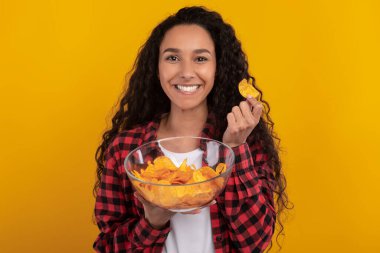Gülümseyen genç Latin kadın canlı sarı-turuncu bir duvara poz verirken elinde bir kase patates cipsi tutuyor. Neşeli bir anda atıştırmanın hazzını yakalamış gibi görünüyor..
