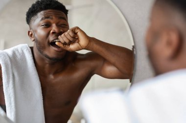 Sabahları, genç siyah bir adam modern bir banyoda üstsüz duruyor. Esniyor ve yumruğunu ağzının yanında tutuyor yansımasına bakarken, güzellik rutinine başlıyor..