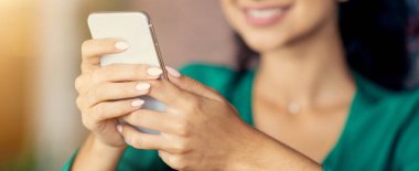 Genç bir kadın akıllı telefonunu tutarken gülümsüyor. Bir kafede oturuyor, görünüşe göre eğleniyor. Doğal ışık yumuşak bir şekilde parlar ve sıcak bir atmosfer yaratır..