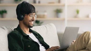 Genç bir adam kanepeye rahatça oturur, kulaklık takar ve dizüstü bilgisayarını kullanırken gülümser. Konforlu oturma odasında bitkiler ve dinlenmek için mükemmel sıcak bir atmosfer var..