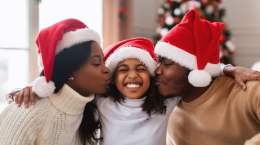 Noel Baba şapkası takmış neşeli siyah aile evde sevgi dolu bir anı paylaşıyor. Ebeveynler birlikte Noel 'i kutlarken gülümseyen kızlarını öperler. Sahne rahat ve şenlikli..