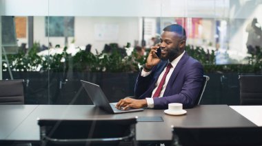 Ofiste dizüstü bilgisayarda çalışan neşeli genç siyahi iş adamı, kişisel asistanı veya iş ortağıyla telefon görüşmesi yapıyor ve gülümsüyor, kopya alanı olan bir panorama, cam duvarlardan vurularak öldürülmüş.