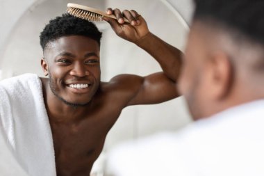 Neşeli siyah bir adam modern bir banyoda üstsüz duruyor, kısa saçlarını tarıyor. Aynadaki yansımasına gülümsüyor, sabah rutinini kucaklıyor..