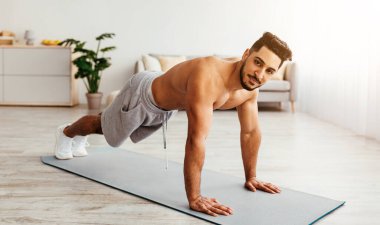 Genç bir adam, modern bir oturma odasında spor minderinde şınav çekiyor. Egzersiz yaparken, spor yaparken, spor yaparken ve sağlığa olan bağlılığını gösterirken güçlü bir formda kalıyor..