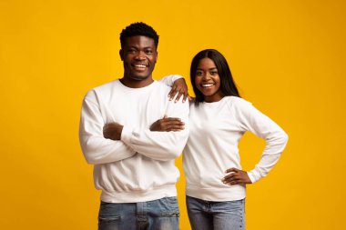 Genç bir çift kameraya gülümseyerek birlikte mutlu bir şekilde duruyorlar. Beyaz sweatshirt ve mavi kot pantolon giyiyorlar. Sahneye enerji katan canlı sarı bir arka plan..