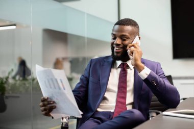 Takım elbiseli gülümseyen siyahi girişimci asistanı ya da iş ortağıyla telefonda konuşuyor, dijital tabletle masada oturuyor, belgelere bakıyor, boşluğu kopyalıyor.
