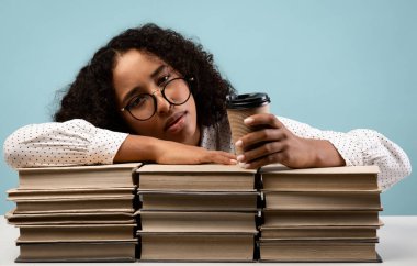 Gözlüklü yorgun siyah bir kız öğrenci kitap yığınlarının üzerine uzanmış, elinde hazır kahveyle bekliyor. Bir stüdyo ortamında yaklaşan üniversite sınavına hazırlanırken yorgun görünüyor..