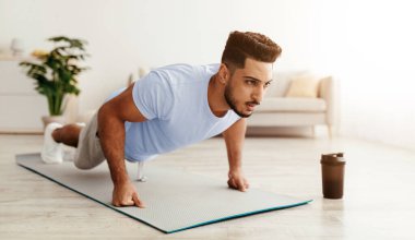 A man performs push-ups on a yoga mat in a bright and airy living room. The scene shows a blend of fitness and relaxation, with a coffee cup nearby, suggesting a morning routine.