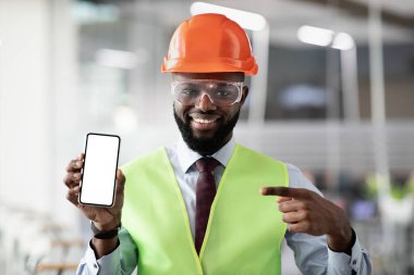 Güvenlik önlemi ve kaskında akıllı telefon taşıyan, beyaz beyaz ekran aletini işaret eden, gülümseyen, taklit eden, profesyonel ihtiyaçlar için mobil uygulama öneren Afro-Amerikan genç erkek mimar.