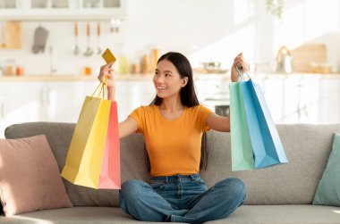 Genç Asyalı bir kadın renkli alışveriş torbalarıyla çevrili bir kanepede oturuyor. Bir kredi kartı tutuyor, son online alışverişleri için heyecanlanıyor ve rahatlatıcı bir anın tadını çıkarıyor..