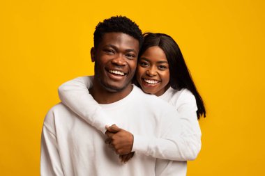 Neşeli bir çift birlikte poz verir, kadın adama arkadan sarılır. İkisi de beyaz gömlek giyer ve geniş bir gülümseme sergilerler. Parlak sarı bir arkaplan önünde mutluluğu yayarlar..