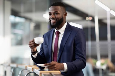 Yakışıklı sakallı Afro-Amerikan iş adamı ofisinde kahve molası veriyor, ofisinin önünde elinde kahveyle duruyor, fotokopi odasına bakıyor ve gülümsüyor.