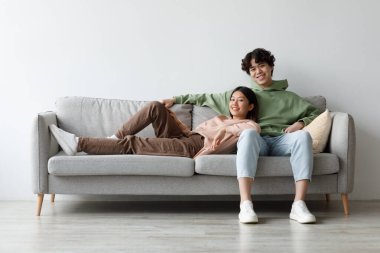 Milenyum Asyalı çiftler evlerindeki kanepede sarılıp rahatlatıcı bir hafta sonunun tadını çıkarırken gülümsüyorlar. Samimi yaşam alanlarında bir anlık mutluluk ve rahatlık paylaşıyorlar..