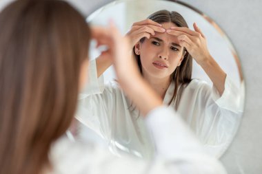 Genç bir kadın banyoda aynanın karşısında duruyor, yüzünü kontrol ederken kaşlarını çatıyor ve alnına sivilce çıkarmaya çalışıyor. Sivilcesi yüzünden üzgün görünüyor..