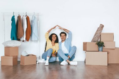 Genç bir çift boş bir odanın zemininde otururken gülümsüyor. Elleriyle bir kalp şekli yaratırlar, raftaki kutular ve kıyafetlerle çevrili..