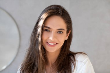 Pürüzsüz tenli ve gür saçlı genç bir bayan evdeki parlak banyoda kameraya bakarken gülümsüyor. Doğal güzelliğini takdir ederek kendine güven ve mutluluk yayıyor..