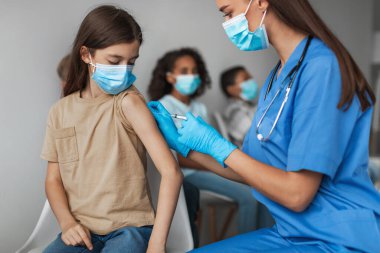 Genç bir kız klinik ortamında Covid-19 'a karşı aşı oluyor. Bir sağlık çalışanı aşıyı yönetirken diğer çocuklar da güvenlik için maske takıyor..
