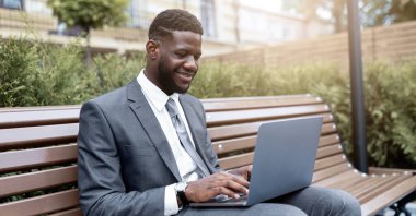Takım elbiseli bir adam bir bankta oturuyor, dizüstü bilgisayarıyla gülümsüyor. Yeşil bitkiler onu parlak ve güneşli bir yerde çevreleyip hoş bir çalışma ortamı yaratıyor..