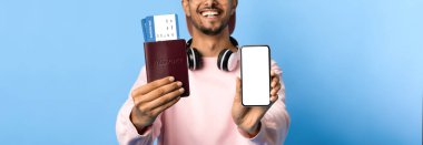 Genç bir adam bir elinde akıllı telefon, diğer elinde seyahat biletleri olan bir pasaport sergilerken gülümsüyor. Kulaklık takıyor ve parlak mavi bir arkaplanı var..