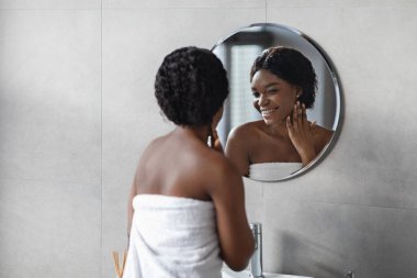 Beyaz bir havluya sarılmış genç siyah kadın aynada kendine hayran, duştan sonra pürüzsüz tenini hissediyor. Parlak bir banyo ortamında zevkle boynuna dokunur..