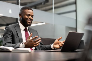 Karizmatik genç siyah yönetici ortaklarıyla iş görüşmesi yapıyor, modern ofiste çalışma masasında oturuyor, laptop kullanıyor, bilgisayar ekranına bakıyor, jest yapıyor ve gülümsüyor, boşluğu kopyalıyor.