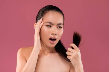 At kuyruklu çekici Asyalı kadın saçında kırık var mı diye incelerken sıkıntılı görünüyor. Kafasına dokunuyor ve minimalist mor bir stüdyo ortamında endişe gösteriyor..