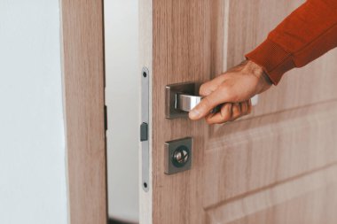 Bir insan elini uzatır ve parlak bir iç mekanda kapı kolunu tutar. Kapı modern bir tasarımı gösteriyor. Ahşap kaplama, günlük aktiviteleri gösteriyor..