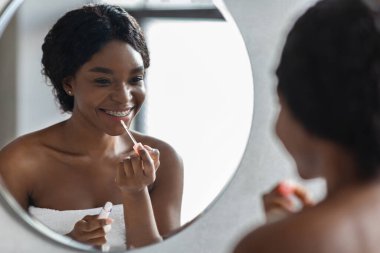 Gülümseyen genç siyah bir kadın aynaya bakarken dudak parlatıcısı sürüyor. Sabah duşunu yeni bitirdi ve dudaklarını gri bir banyoda nemlendiriyor..