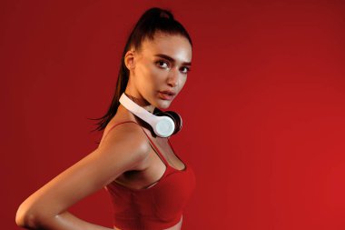 Bir fitness modeli parlak kırmızı bir ortamda kendine güvenen bir poz verirken spor kıyafeti ve kulaklık takar. Işıklandırma onun özelliklerini geliştiriyor, çarpıcı bir görsel cazibe yaratıyor..