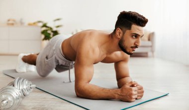 A man with an athletic build is holding a plank position on a yoga mat inside a well-lit room. Dumbbells are positioned nearby, emphasizing his workout regimen.