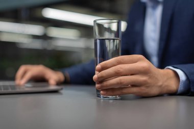 Takım elbiseli tanınmayan bir yönetici bir masada oturuyor, elinde bir bardak su tutarken dizüstü bilgisayarda yazıyor. Ofisin içi parlak ve profesyoneldir, geniş bir kopya alanı vardır..