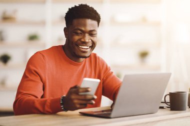 Bir adam ahşap bir masada otururken gülümsüyor, bir elinde akıllı bir telefon ve önünde bir dizüstü bilgisayar kullanıyor. Parlak atmosfer rahat bir çalışma ortamı olduğunu gösteriyor..