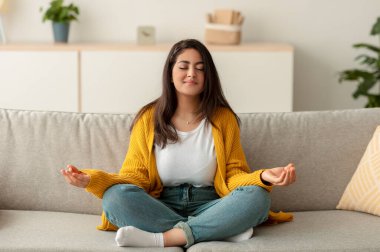 Huzurlu bir Arap kadın kanepesinde nilüfer pozisyonunda oturur ve gözleri kapalı meditasyon yapar. Evde sakin bir anın tadını çıkarıyor, hafta sonu boyunca dinlenmeye odaklanıyor..