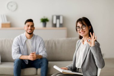 Genç bir kadın psikolog başarılı bir terapi seansı sırasında bin yıllık bir Arap 'a rehberlik ederken iyi bir işaret gösteriyor. Çağdaş bir ofiste, destekleyici bir atmosfer oluşturuyorlar..