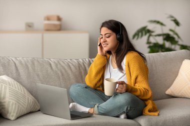 Ortadoğulu kadın kanepesinde rahatça oturur ve uzaktan kumandayla meşgul olur. Kulaklık takıyor, elinde bir fincan kahve tutuyor ve evde verimli bir seans sırasında bilgisayarına odaklanıyor..