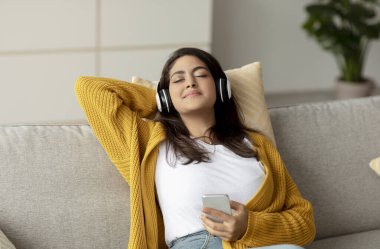 Genç, rahat bir Arap kadın rahat bir koltukta kablosuz kulaklık takıyor. O, akıllı telefonundan müzik dinler, en sevdiği şarkıları ve evdeki huzurlu ortamı sever..