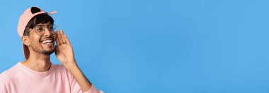 Gözlüklü genç bir adam sevinçle ellerini ağzına koyuyor, sanki haberleri paylaşıyor ya da başkalarına sesleniyormuş gibi. Pembe bir şapka ve süveter giyer, parlak mavi bir arka planda durur..