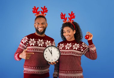 Yeni yılı kutluyoruz. Gece yarısına beş dakika kala, geyik boynuzu ve süveter giyen, mavi arka planda poz veren mutlu siyah eşleri olan stüdyo çekimi.