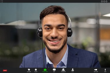 Genç ve mutlu bir Orta Doğulu işadamı müşterileriyle bir video görüşmesi yapıyor, gülümsüyor ve doğrudan kameraya bakıyor..