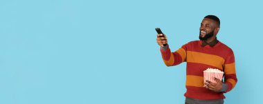 Heyecanlı genç siyahi adam televizyon izliyor ve patlamış mısır yiyor. Afro-Amerikan adam elinde uzaktan kumandayla kanal değiştiriyor. Mavi arka planda, panoramada, kopyalama alanında tek başına duruyor.