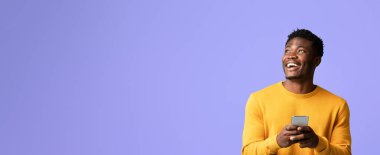 Bir adam elinde akıllı bir telefonla gülümsüyor, sevinç belirtileri gösteriyor. Parlak mor bir arka plana karşı duruyor, neşeli bir atmosfer yaratıyor..