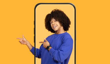 Kıvırcık saçlı genç bir birey şakacı bir şekilde poz veriyor ve bir telefon çerçevesi içinden işaret ediyor. Parlak arka plan neşe katar, bir etkileşim ve eğlence anı yakalar..