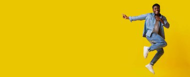 Bir adam büyük bir gülümsemeyle sevinçle havada zıplıyor. Sıradan bir kot pantolon ve kot gömlek giyiyor. Parlak sarı arka plan, o anın canlı atmosferini arttırır..