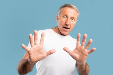 Olgun bir adam korkmuş görünüyor, doğrudan kameraya bakıyor. Korkuyu ifade ederek ve kendini savunmak için elini kaldırdı. Mavi bir stüdyonun arka planına karşı durdu..