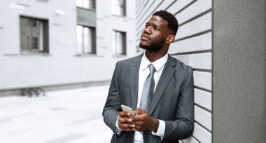 Takım elbiseli bir işadamı modern bir binanın önünde duruyor ve akıllı telefonuna dikkatlice bakıyor. Kentsel ortam gün içinde profesyonel bir atmosfer yaratıyor..