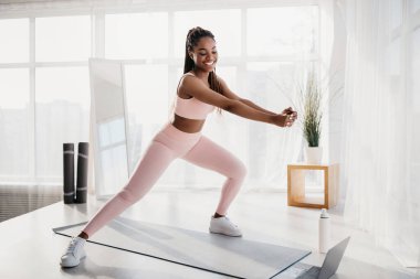 Genç Afrikalı Amerikalı kadın, laptopundaki bir spor videosunun ardından kapalı alanda bir paspasa atlıyor. İyi ışıklandırılmış evinde aktif bir yaşam tarzını seviyor, spor rutinine odaklanıyor..